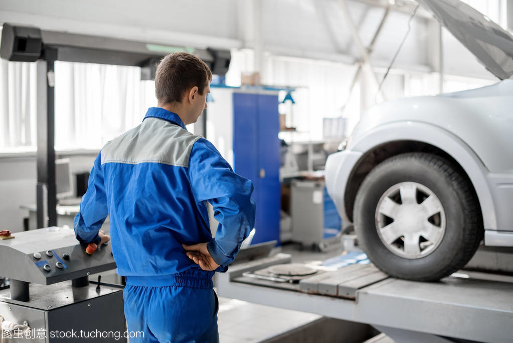 一个在一个蓝色工作形式的汽车车间工作的技工正在修理一辆灰色的汽车。汽车服务